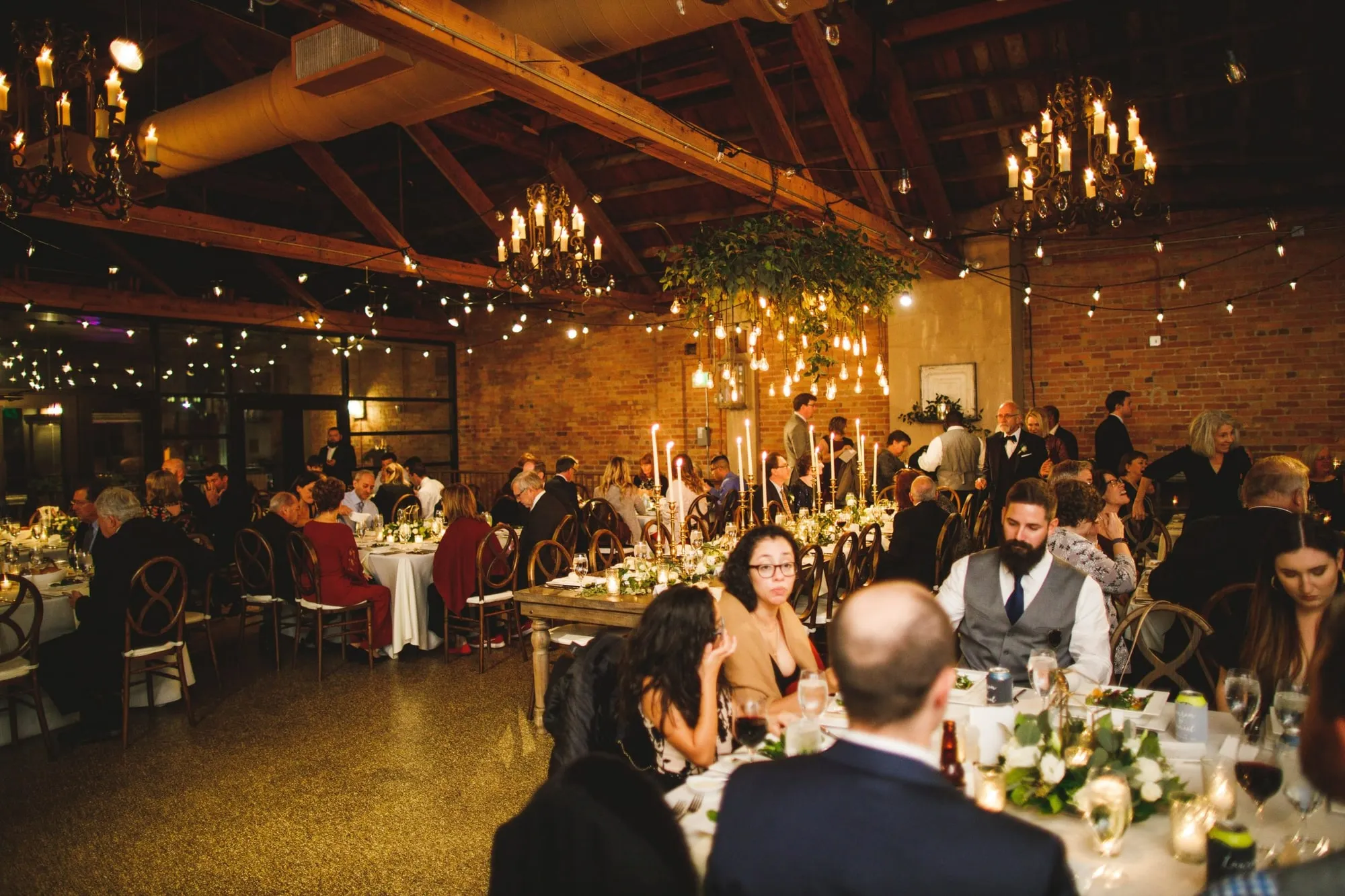 A dinner underway by candlelight, tall tapers down the center of the tables