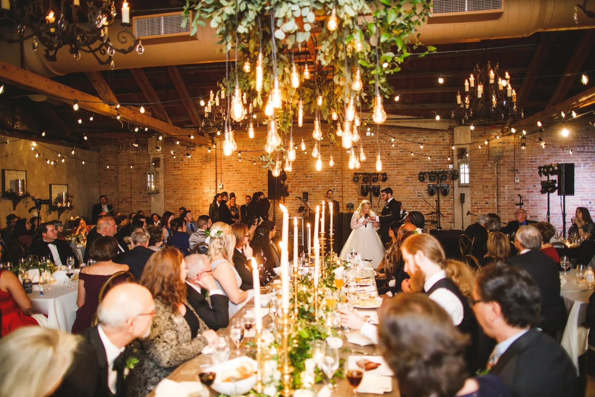 Bare filament bulbs hung at varying heights above a long table, greenery threaded between them