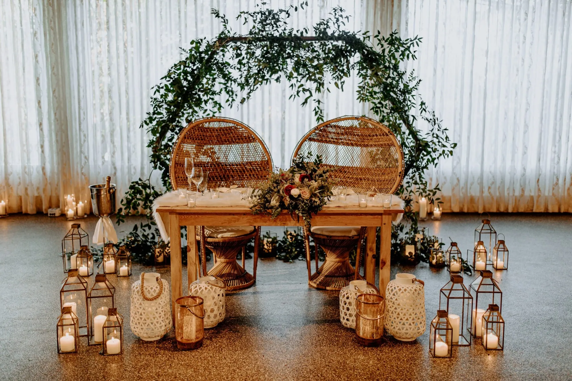 A sweetheart table with two rattan chairs beneath an arch of greenery, lanterns at the base