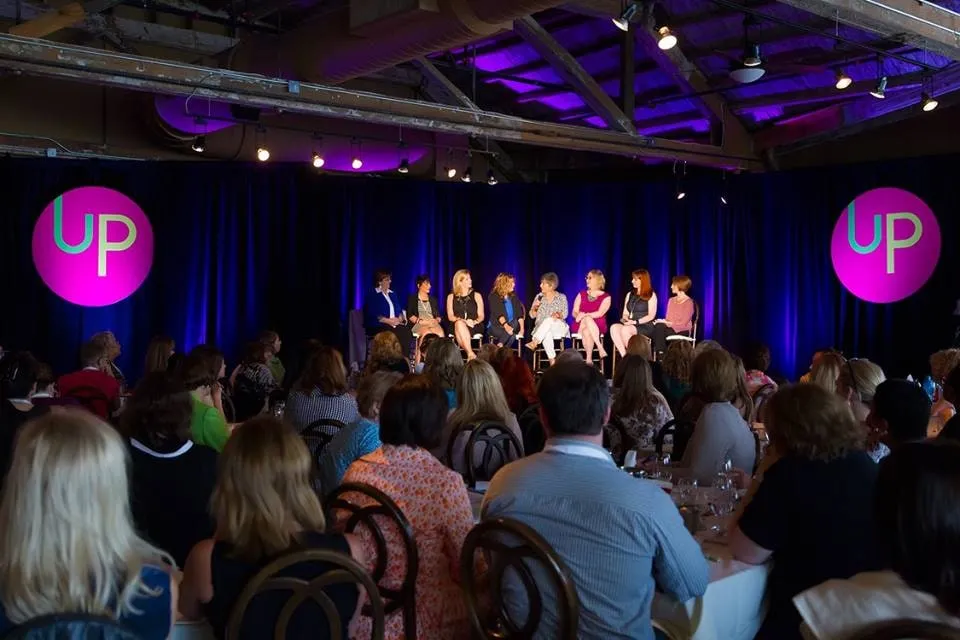 A panel of speakers on a lit stage in front of a full seated audience
