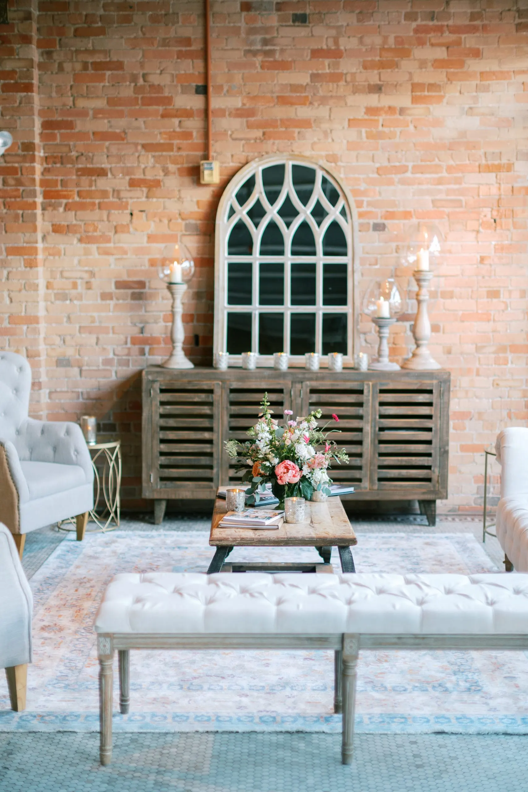 A lounge against the exposed brick: tufted armchairs and a bench around a low wooden table, an arched mirror and candles on the sideboard behind