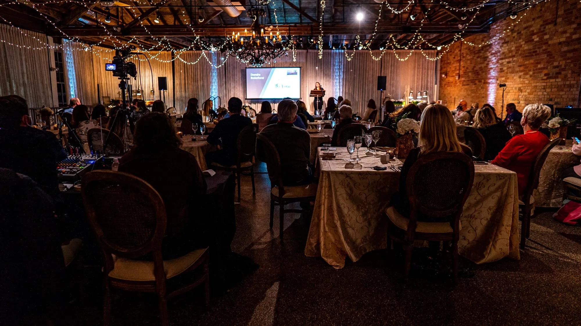 A seated corporate dinner in progress, a speaker at a lit podium beside a projection screen